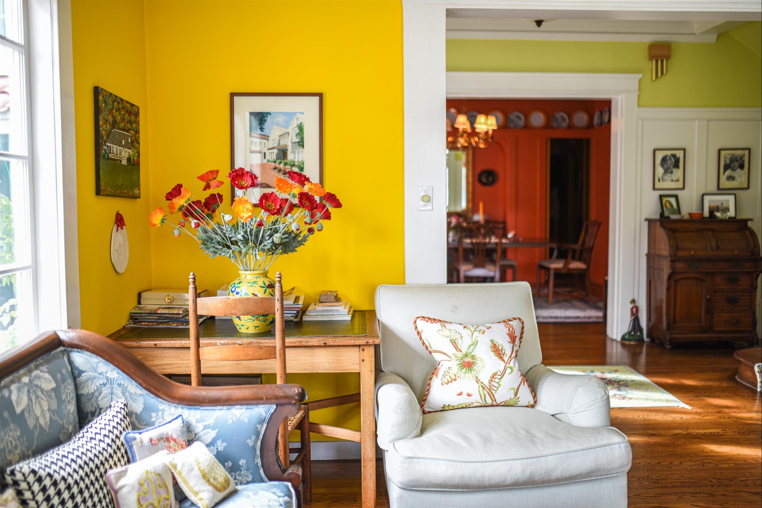 A brightly lit living room with a yellow wall, a wooden desk, and a floral vase.